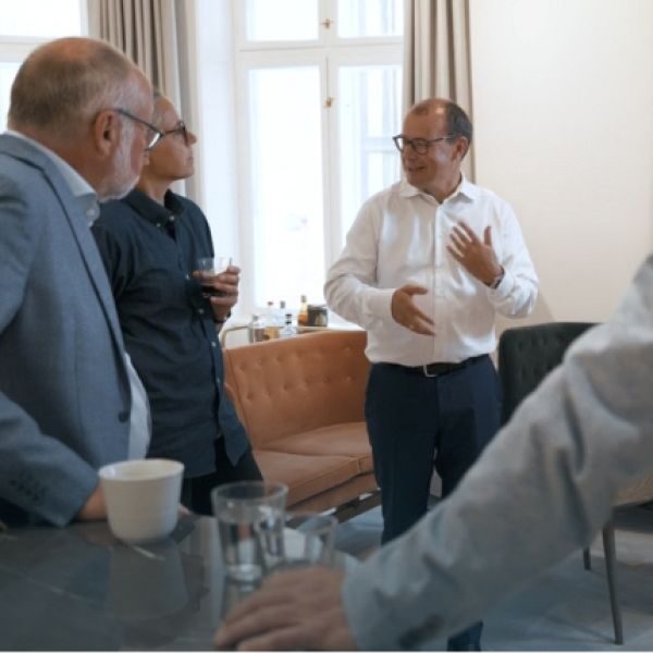 En gruppe mænd i samtale i et lyst mødelokale med sofa og kaffekopper på bordet.
