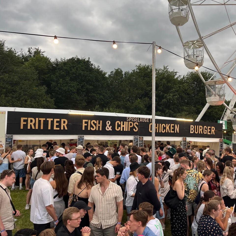 Stor menneskemængde foran madbod med skilte for ‘Fritter’, ‘Fish & Chips’, ‘Chicken’ og ‘Burger’ på en udendørs festival med pariserhjul i baggrunden.